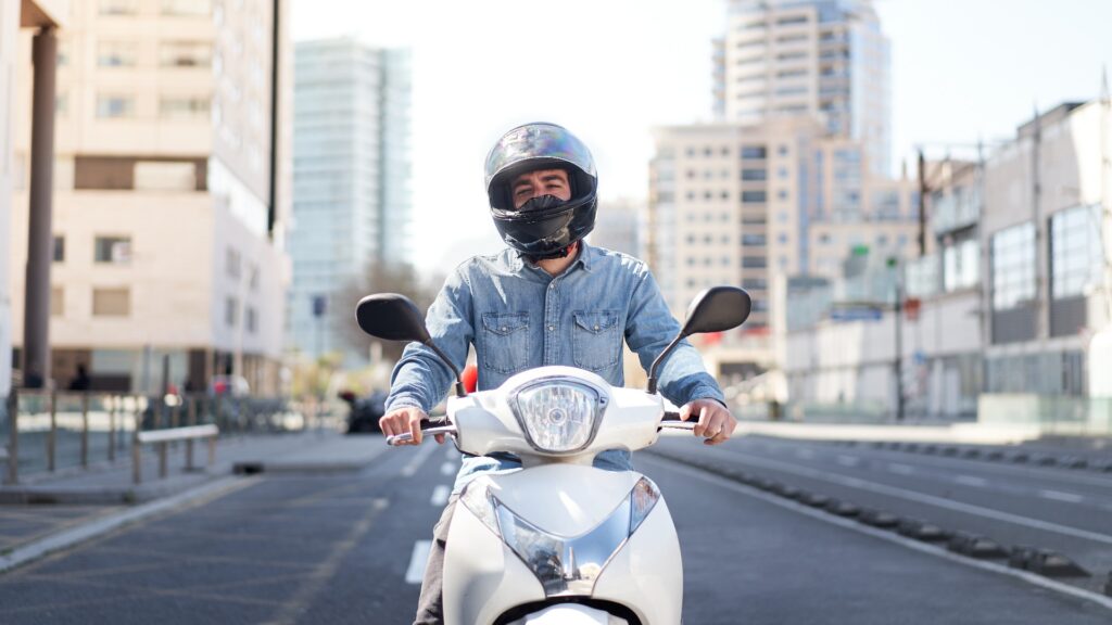 Mittlere Einstellung eines jungen Motorradfahrers, der an einer Ampel in Barcelona steht. Der Typ fährt mit seinem weißen Roller durch die Stadt auf einer großen Straße voller Wolkenkratzer.