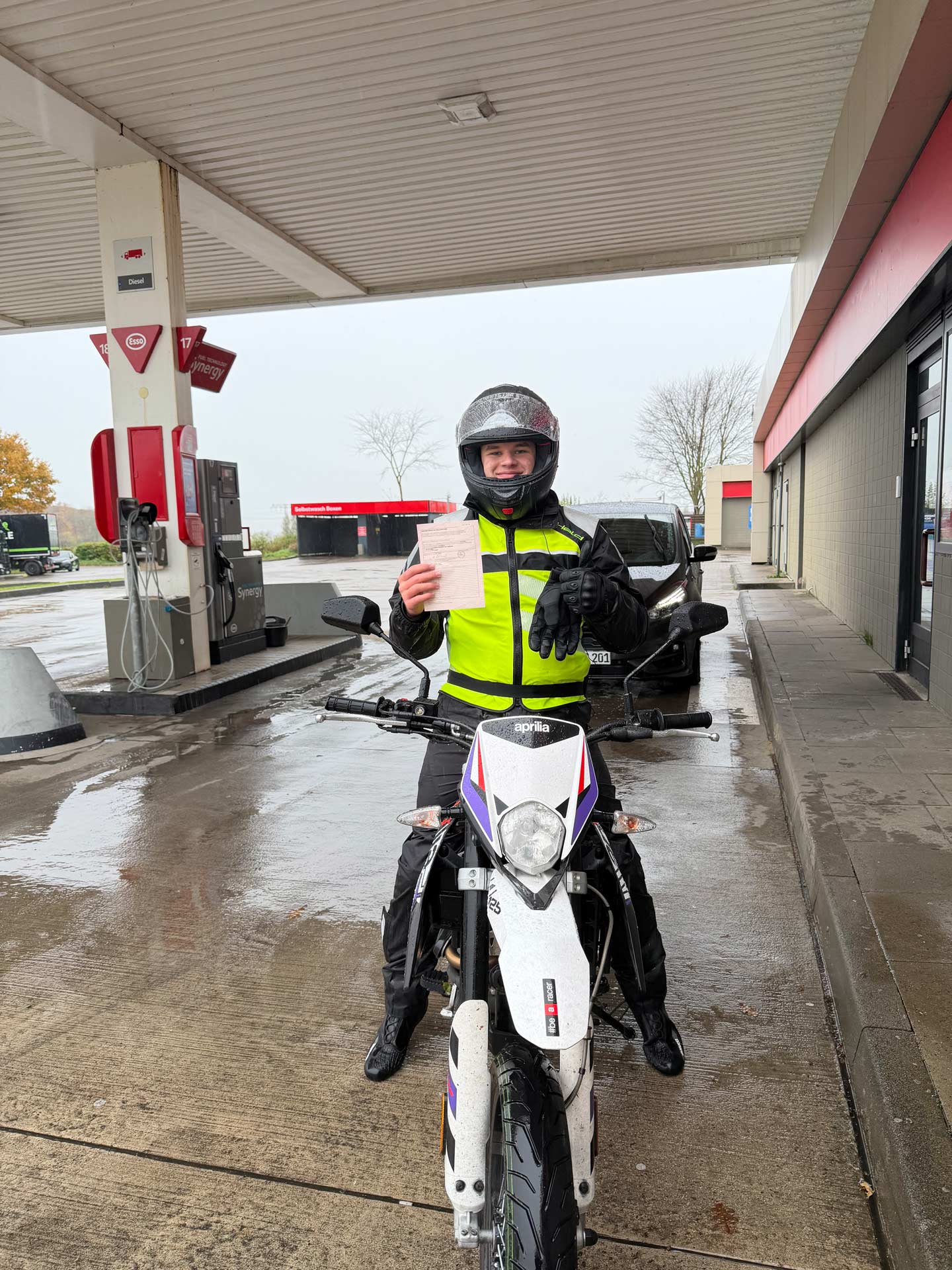 Eine lachende Person im Motorradoutfit hält stolz ein Dokument an einer Tankstelle hoch