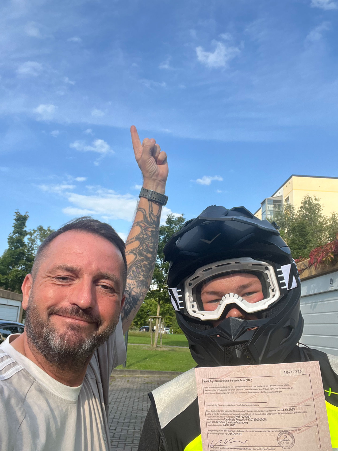 Zwei Männer lächeln, einer mit Helm und Dokument, anderer zeigt lachend zum Himmel