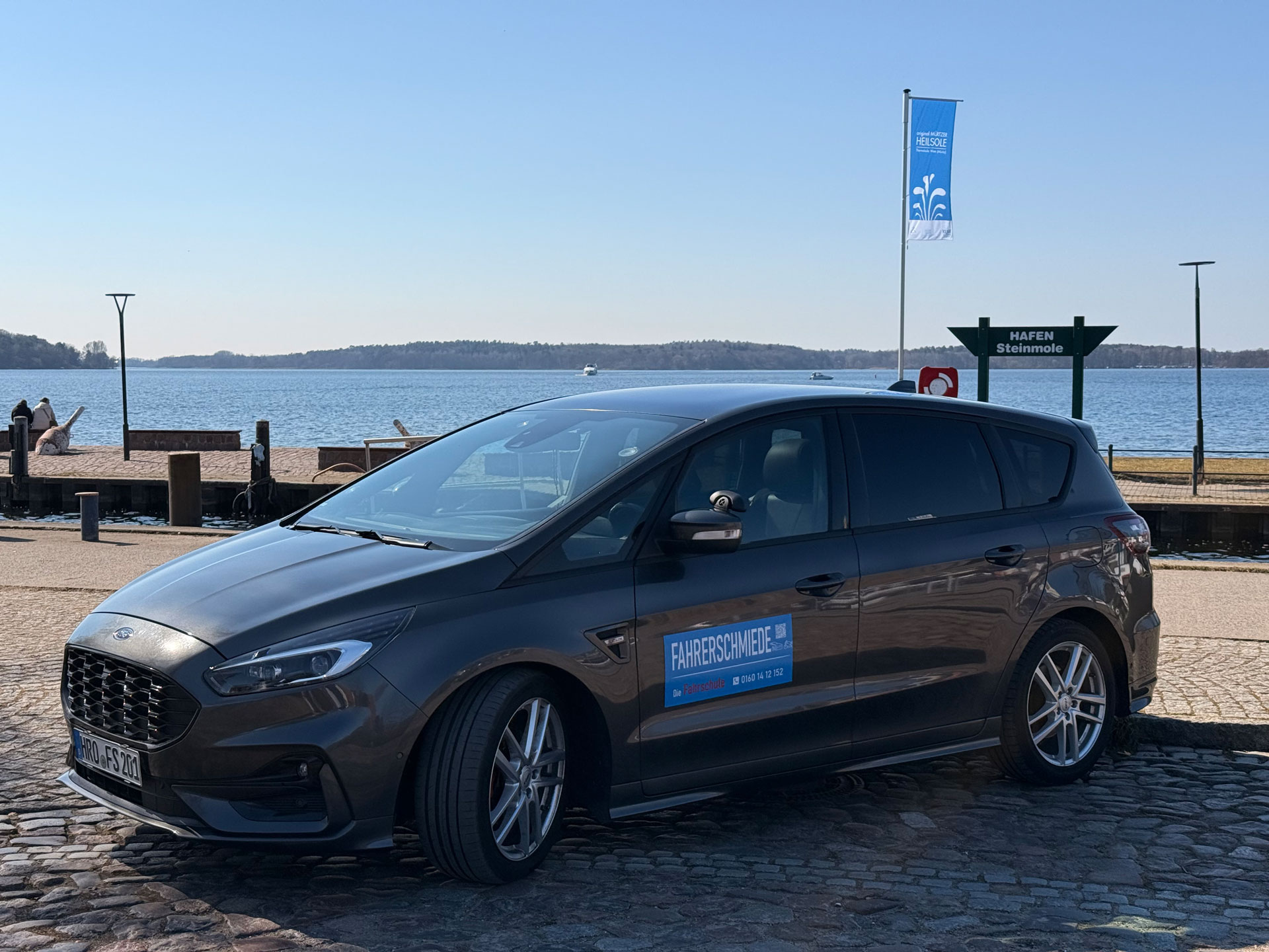 Ein grauer Ford steht auf einem Pflasterweg vor einem See bei sonnigem Wetter