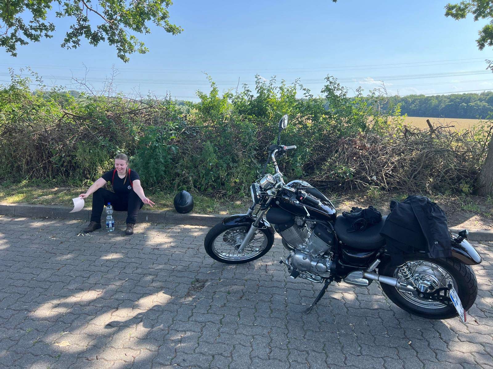 Eine lächelnde Person sitzt entspannt auf dem Boden neben einem schwarzen Motorrad