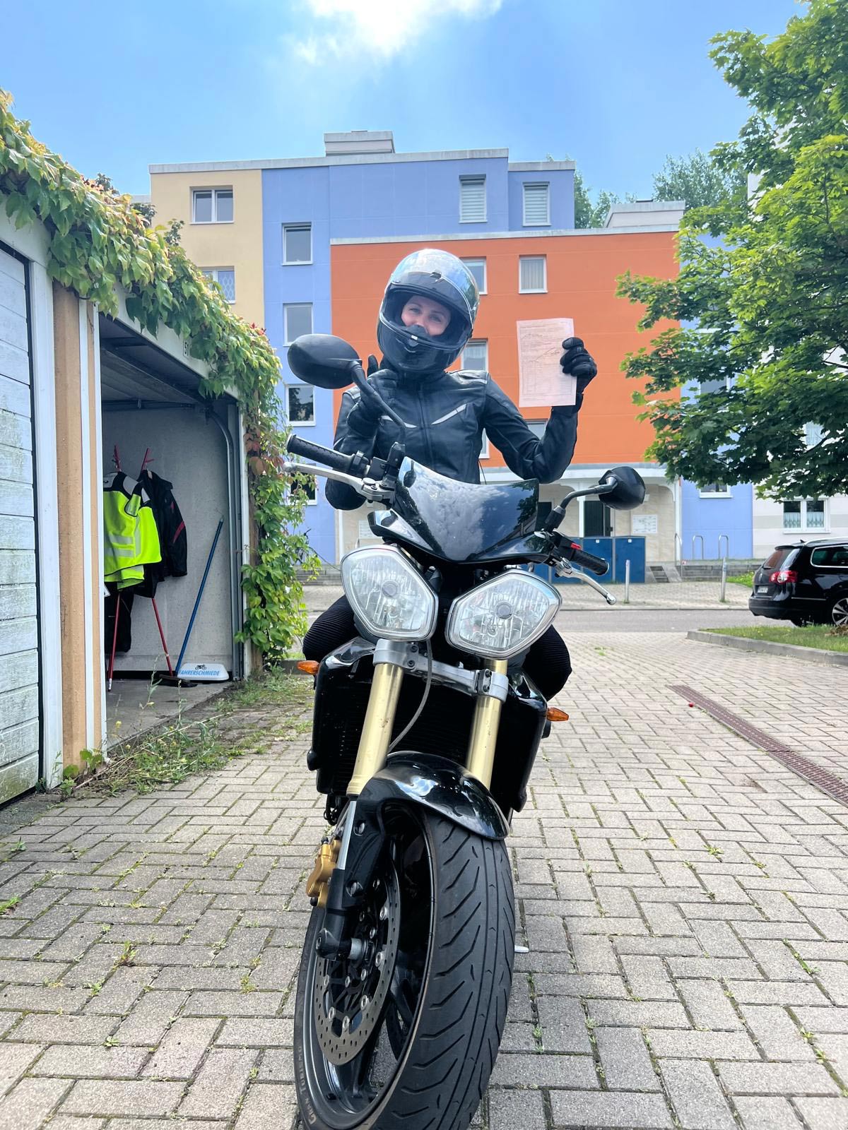 Eine Frau in schwarzer Motorradkleidung und Helm lächelt stolz und hält ein Dokument hoch