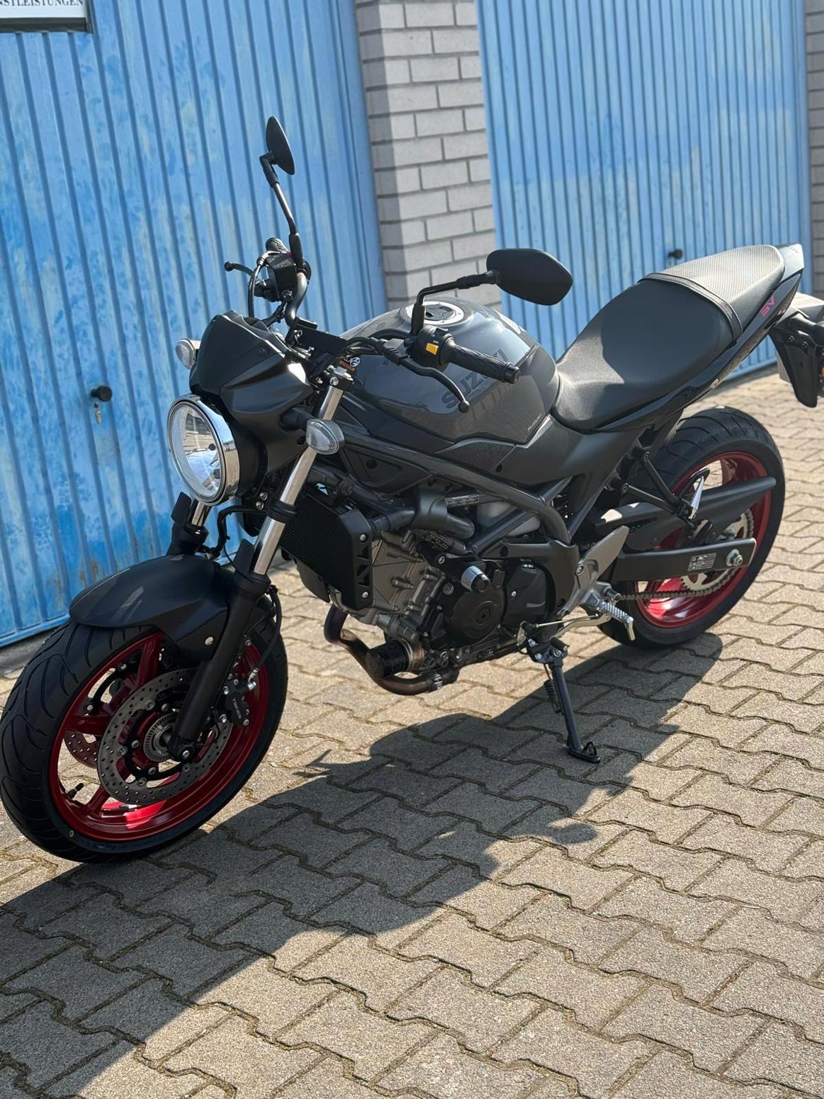 Schwarzes Motorrad mit roten Felgen steht vor einem blauen Garagentor im Sonnenschein